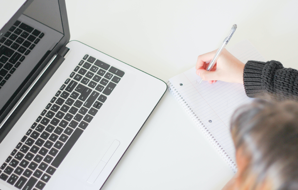 Open laptop with someone writing right handed on a spiral notebook in front of it